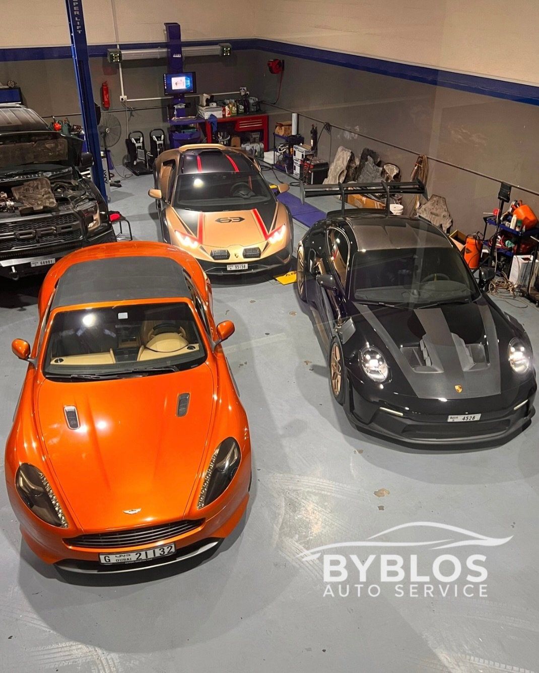 An orange Aston Martin, a black Porsche 911 and a Lamborghini parked on the workshop floor