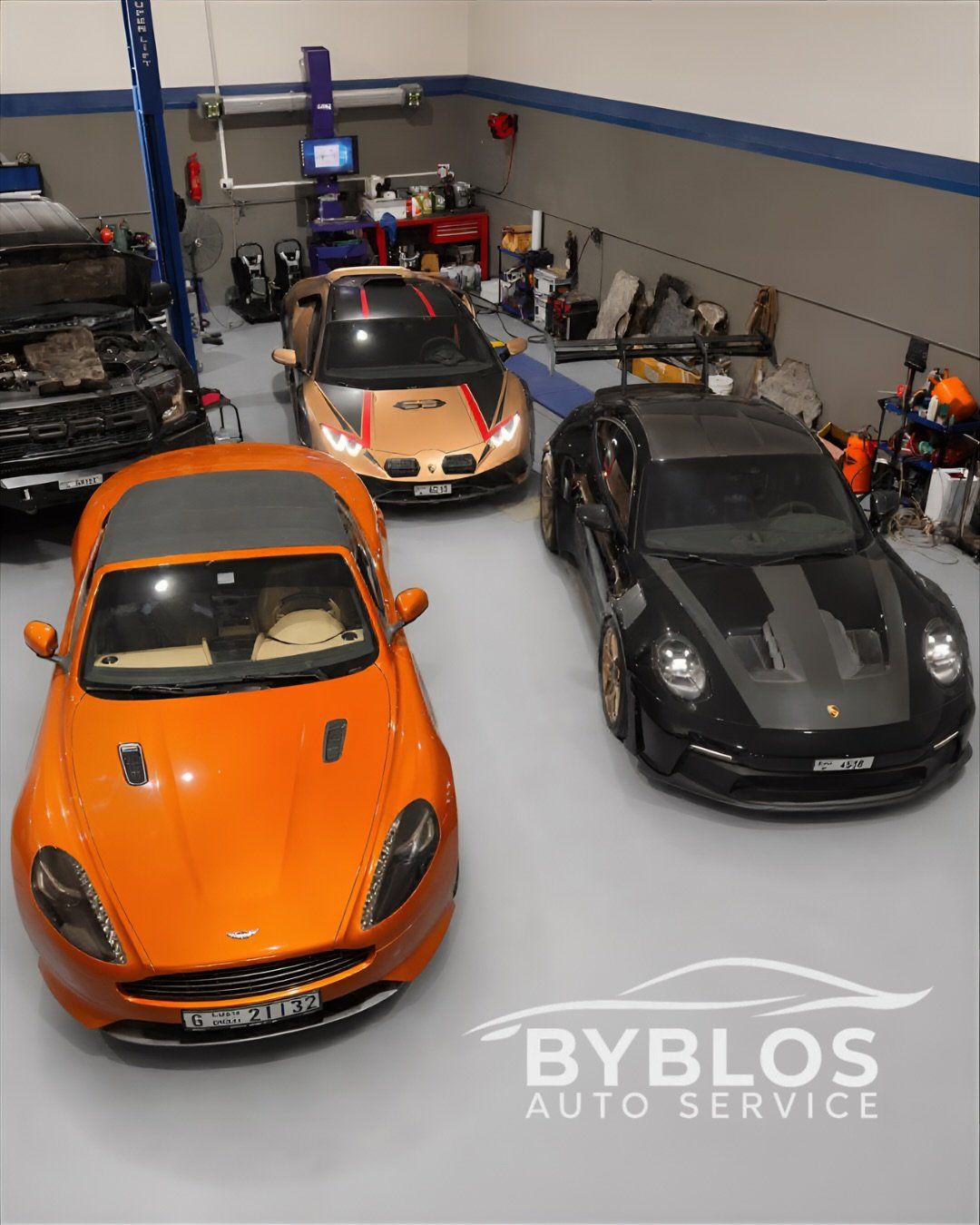 Overhead view of an orange Aston Martin, a black Porsche and a Lamborghini in the BYBLOS workshop